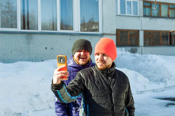 Dos hombres fotografiando themselves.guy con barba, vestido con chaqueta casual deportes de otoño y sombrero naranja brillante, hace selfie en el teléfono en caso elegante, caminando con su amigo al aire libre día de invierno soleado — Foto de Stock