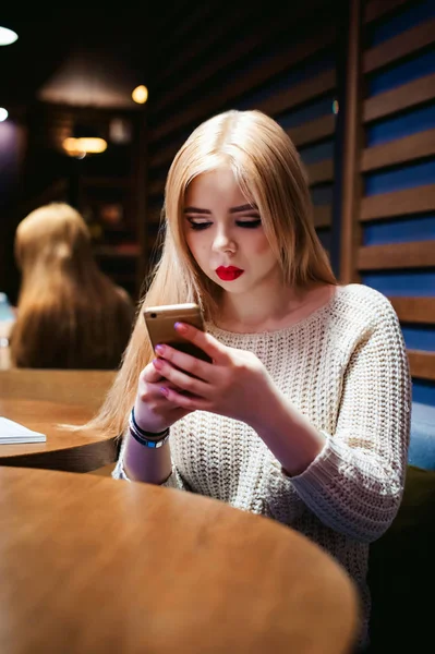 年轻的金发美女在针织毛衣、 自拍照照片制作一部智能手机，坐在一家咖啡馆，坐在餐桌上花时间独处，做互联网通信 — 图库照片