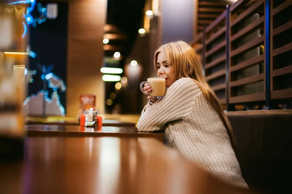 Carino giovane donna bionda beve caffè caldo da una tazza di vetro all'interno di una caffetteria — Foto Stock