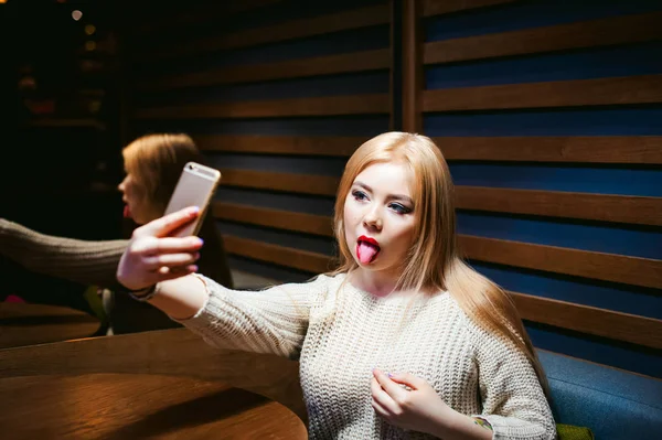 Wanita muda berambut pirang dalam sweater rajutan, membuat foto selfie pada smartphone, duduk di meja di kafe, menghabiskan waktu sendirian, melakukan komunikasi internet Stok Gambar