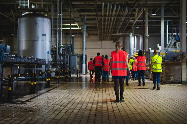 Blonde woman in overalls, on the tour of the brewery plant in industrial premises, amid crowds of people in vests, inspecting a factory for training purposes — Stock Photo, Image