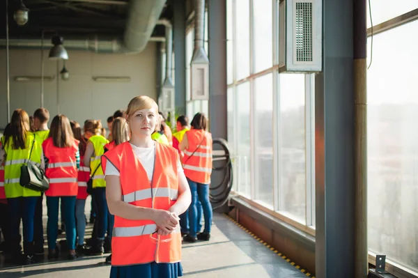 Blonde woman in overalls, on the tour of the brewery plant in industrial premises, amid crowds of people in vests, inspecting a factory for training purposes — Stock Photo, Image