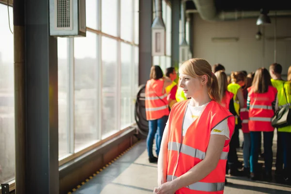 Blonde woman in overalls, on the tour of the brewery plant in industrial premises, amid crowds of people in vests, inspecting a factory for training purposes — Stock Photo, Image