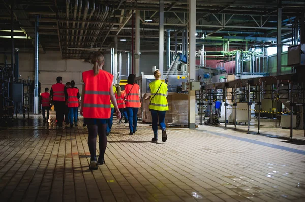 Blonde woman in overalls, on the tour of the brewery plant in industrial premises, amid crowds of people in vests, inspecting a factory for training purposes — Stock Photo, Image
