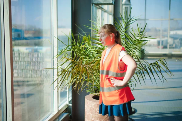 Female employee in orange robe wearing vest in working space production room, against background large windows all way from ceiling to floor. Controls logistic process occurring on territory outside — Stock Photo, Image