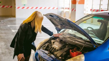 kadın araba hood yakınındaki. Genç sarışın Alışveriş Merkezi, standları ile yükseltilmiş motor bölmesi kukuleta, car yakınındaki kapalı otopark içinde telefon hizmeti tarafından arıza ile sorunu çözdü