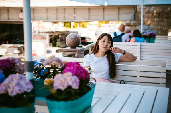 Linda mujer joven asiática en el café de verano al aire libre. chica en camiseta blanca, con el pelo largo en el interior acogedor luz simple del restaurante de estilo urbano — Foto de Stock