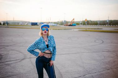 güneş gözlüğü, şapka ve kot pantolon, mavi ekose desenli gömlek giymiş genç ince kadın yaz gününde şafak siyah seyahat bavul ile özgürlük gezisinde kalkacak