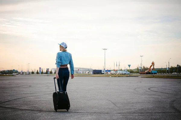 Joven mujer delgada vestida con camisa azul a cuadros Gafas de sol, gorra y jeans, Saldrá al amanecer en el día de verano en viaje de libertad con maleta de viaje negro — Foto de Stock