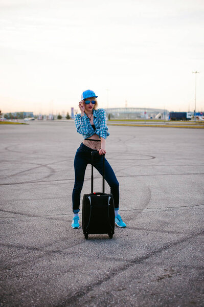 young slim woman dressed in blue checked shirt Sunglasses, cap and jeans, Will depart at dawn on summer day on freedom trip with black travel suitcase