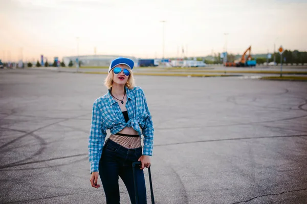 Young slim woman dressed in blue checked shirt Sunglasses, cap and jeans, Will depart at dawn on summer day on freedom trip with black travel suitcase — Stock Photo, Image