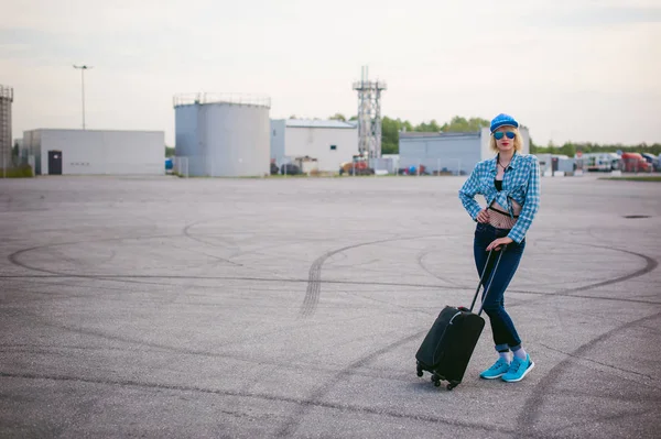 Giovane donna magra vestita in camicia a quadri blu Occhiali da sole, berretto e jeans, partirà all'alba il giorno d'estate in viaggio di libertà con valigia da viaggio nera — Foto Stock