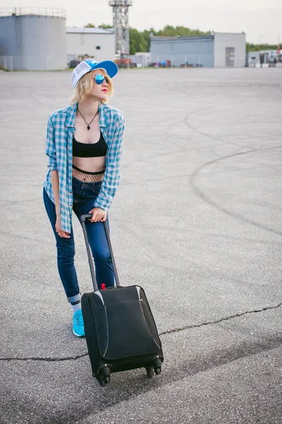 Giovane donna magra vestita in camicia a quadri blu Occhiali da sole, berretto e jeans, partirà all'alba il giorno d'estate in viaggio di libertà con valigia da viaggio nera — Foto Stock