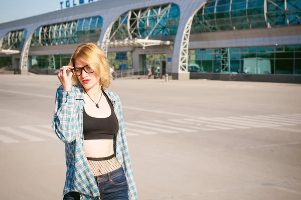 Giovane donna magra vestita in camicia a quadri blu, jeans, occhiali. Partirà all'alba del giorno d'estate in viaggio di libertà con valigia nera contro l'edificio dell'aeroporto — Foto Stock