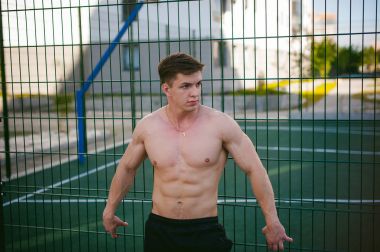 Yakışıklı seksi erkek vücut geliştirmeci sporcu adam çıplak gövde ile bir güçlü kas vücut ayakta gösterilen pozlar courtyard bir yeşil kılavuz çit spor alanında yakınındaki. Sağlıklı yaşam konsepti.