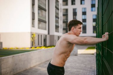 Yakışıklı seksi erkek vücut geliştirmeci sporcu adam çıplak gövde ile bir güçlü kas vücut ayakta gösterilen pozlar courtyard bir yeşil kılavuz çit spor alanında yakınındaki. Sağlıklı yaşam konsepti.