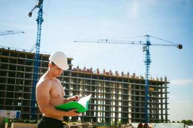 Çıplak bir gövde ile genç seksi erkek vücut geliştirmeci atlet. İnşaat yapı ve inşaat vinç belgeler ile arka plan üzerinde beyaz koruyucu kask Yöneticisi mühendis Oluşturucu standları