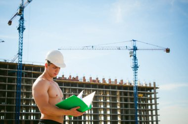 Çıplak bir gövde ile genç seksi erkek vücut geliştirmeci atlet. İnşaat yapı ve inşaat vinç belgeler ile arka plan üzerinde beyaz koruyucu kask Yöneticisi mühendis Oluşturucu standları