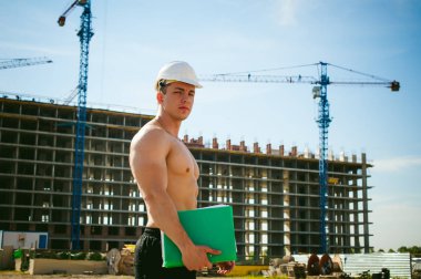 Çıplak bir gövde ile genç seksi erkek vücut geliştirmeci atlet. İnşaat yapı ve inşaat vinç belgeler ile arka plan üzerinde beyaz koruyucu kask Yöneticisi mühendis Oluşturucu standları