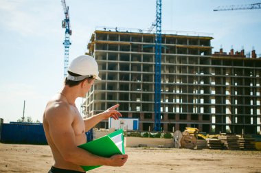 Çıplak bir gövde ile genç seksi erkek vücut geliştirmeci atlet. İnşaat yapı ve inşaat vinç belgeler ile arka plan üzerinde beyaz koruyucu kask Yöneticisi mühendis Oluşturucu standları