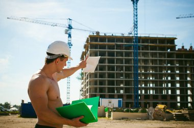 Çıplak bir gövde ile genç seksi erkek vücut geliştirmeci atlet. İnşaat yapı ve inşaat vinç belgeler ile arka plan üzerinde beyaz koruyucu kask Yöneticisi mühendis Oluşturucu standları