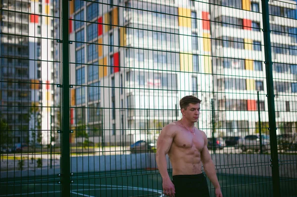 Όμορφος σέξι αρσενικό bodybuilder άνδρας αθλητής με γυμνό κορμό, πόζες που δείχνει ένα ισχυρό μυώδες σώμα στέκεται κοντά σε ένα πράσινο Πλέγμα περίφραξης αθλητικό τομέα στην αυλή. Έννοια του υγιεινού τρόπου ζωής. — Φωτογραφία Αρχείου