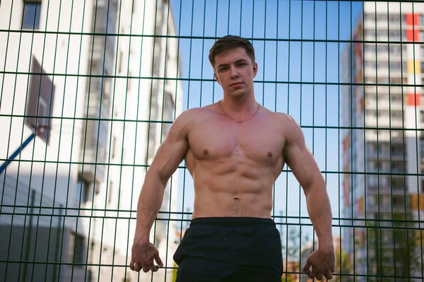 帅气性感的男性健美运动员人与半裸，显示出强大的肌肉发达的身体站立的姿势附近的绿色网格围栏的运动场上，在院子里。健康的生活方式的概念. — 图库照片