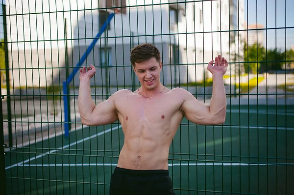 Bello sexy maschio bodybuilder atleta uomo con busto nudo, posa mostrando un potente corpo muscolare in piedi vicino a un campo sportivo recinto griglia verde in cortile. Stile di vita sano concetto . — Foto Stock