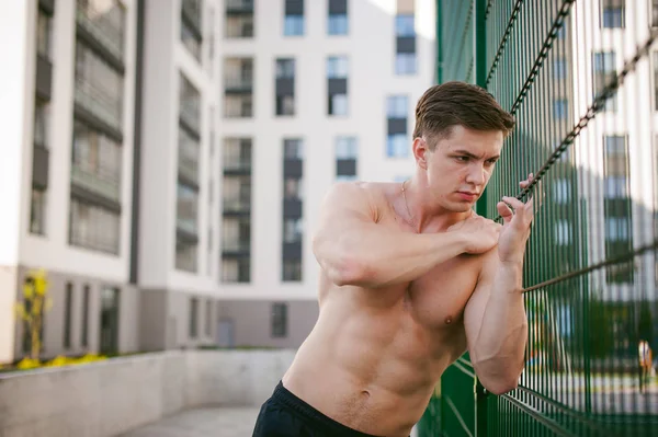 帅气性感的男性健美运动员人与半裸，显示出强大的肌肉发达的身体站立的姿势附近的绿色网格围栏的运动场上，在院子里。健康的生活方式的概念. — 图库照片