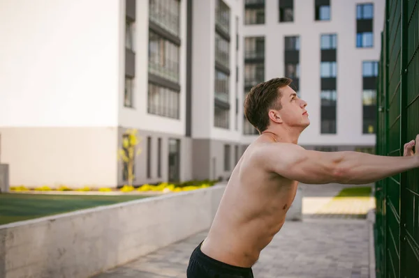 裸の胴体を持つハンサムなセクシーな男性のボディービルダー選手男、強力な筋肉のついた体立っているを示すポーズ近く中庭でグリーン ・ グリッド フェンス スポーツ フィールドです。健康的なライフ スタイルのコンセプト. — ストック写真