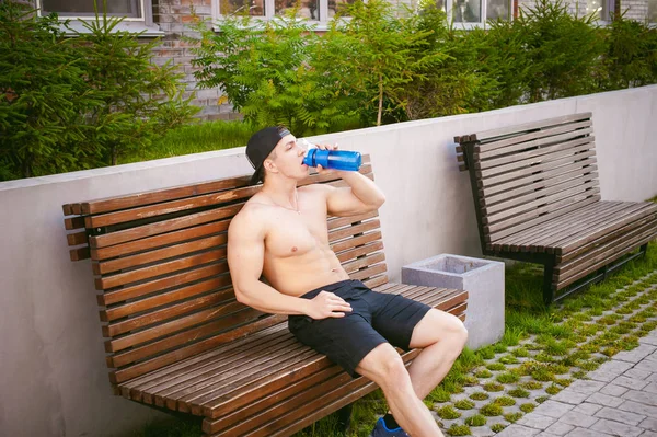 Schöner sexy männlicher Athlet Mann mit nacktem Oberkörper auf einer Bank im Hof des Hauses ruht, nach dem Training trinkt Wasser aus einer Flasche, ein früher sonniger Sommermorgen — Stockfoto