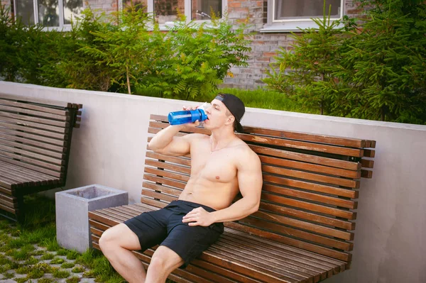 Schöner sexy männlicher Athlet Mann mit nacktem Oberkörper auf einer Bank im Hof des Hauses ruht, nach dem Training trinkt Wasser aus einer Flasche, ein früher sonniger Sommermorgen — Stockfoto