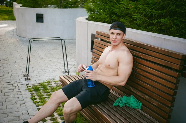 Schöner sexy männlicher Athlet Mann mit nacktem Oberkörper auf einer Bank im Hof des Hauses ruht, nach dem Training trinkt Wasser aus einer Flasche, ein früher sonniger Sommermorgen — Stockfoto