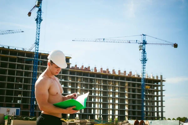 Çıplak bir gövde ile genç seksi erkek vücut geliştirmeci atlet. İnşaat yapı ve inşaat vinç belgeler ile arka plan üzerinde beyaz koruyucu kask Yöneticisi mühendis Oluşturucu standları