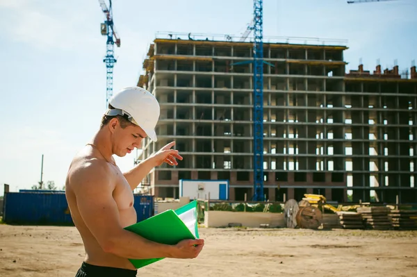 Çıplak bir gövde ile genç seksi erkek vücut geliştirmeci atlet. İnşaat yapı ve inşaat vinç belgeler ile arka plan üzerinde beyaz koruyucu kask Yöneticisi mühendis Oluşturucu standları