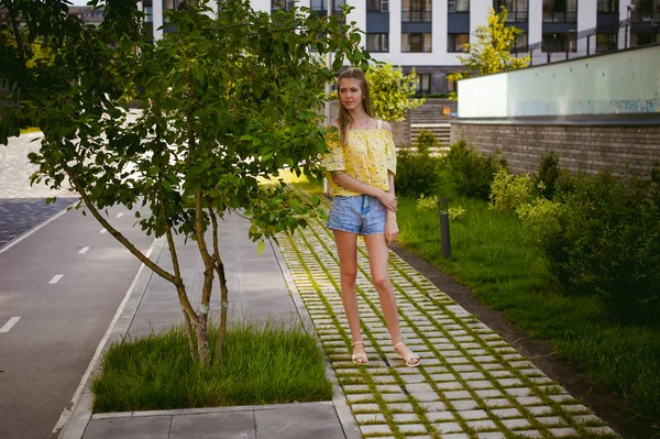 Mujer hermosa joven está paseando al aire libre en el patio, contra el telón de fondo de vegetación y casas, cálido día soleado de verano — Foto de Stock
