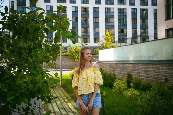 Mujer hermosa joven está paseando al aire libre en el patio, contra el telón de fondo de vegetación y casas, cálido día soleado de verano — Foto de Stock