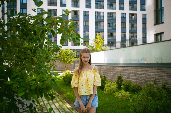 Mujer hermosa joven está paseando al aire libre en el patio, contra el telón de fondo de vegetación y casas, cálido día soleado de verano — Foto de Stock