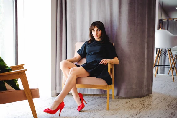 Young attractive woman with long legs in black elegant dress, sits in chair near window in interior of room. Seductive shoes of red light, on beautiful legs