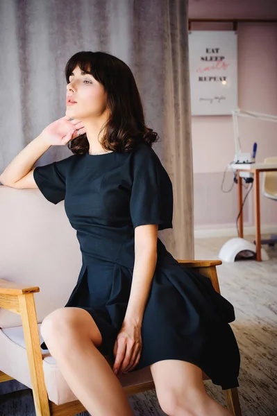 Mujer atractiva joven con piernas largas en vestido elegante negro, se sienta en la silla cerca de la ventana en el interior de la habitación. Zapatos seductores de luz roja, sobre hermosas piernas — Foto de Stock