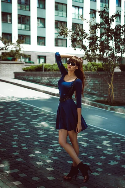 Junge attraktive Frau mit langen Beinen und langen Haaren in einem blau-eleganten Kleid mit einem Schwertgürtel in Sonnenbrille, schlendert durch einen modernen europäischen Hof, ein warmer Sommertag — Stockfoto