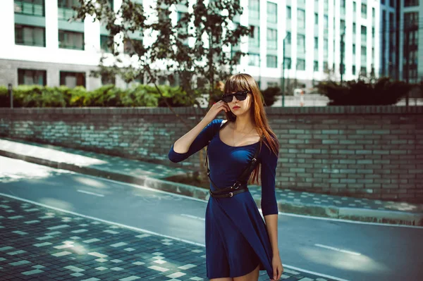 Junge attraktive Frau mit langen Beinen und langen Haaren in einem blau-eleganten Kleid mit einem Schwertgürtel in Sonnenbrille, schlendert durch einen modernen europäischen Hof, ein warmer Sommertag — Stockfoto