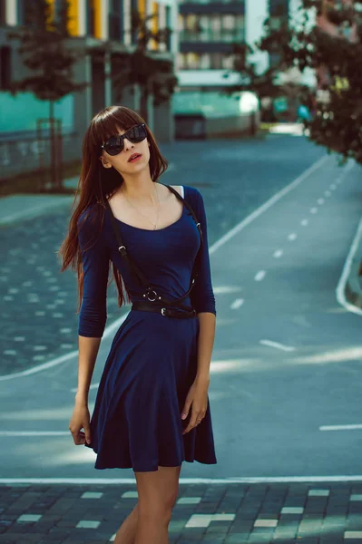 Giovane donna attraente con le gambe lunghe con i capelli lunghi in un abito elegante blu con una cintura di spada In occhiali da sole, passeggiate in un cortile moderno europeo, una calda giornata estiva — Foto Stock