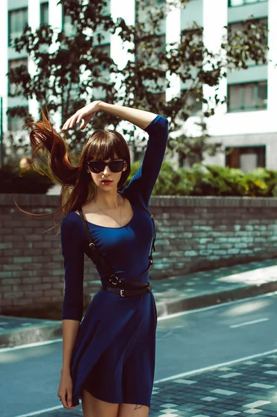 Güneş gözlüğü, bir kılıç kemer ile mavi şık elbiseli uzun saçlı uzun bacaklı genç çekici kadın elini kolunu sallaya dönüyor Avrupa modern avluda bir sıcak yaz günü