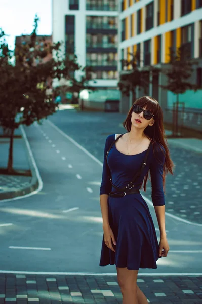 Junge attraktive Frau mit langen Beinen und langen Haaren in einem blau-eleganten Kleid mit einem Schwertgürtel in Sonnenbrille, schlendert durch einen modernen europäischen Hof, ein warmer Sommertag — Stockfoto