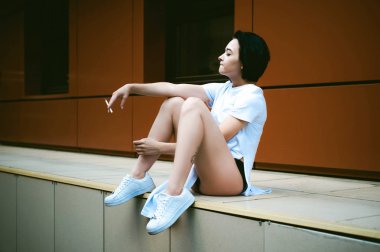 çıplak ayak beyaz spor ayakkabı ve uzun bir T-shirt giymiş genç kadınla cityscapes zemin karşı bir sigara içiyor. Sokak moda