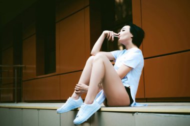 çıplak ayak beyaz spor ayakkabı ve uzun bir T-shirt giymiş genç kadınla cityscapes zemin karşı bir sigara içiyor. Sokak moda