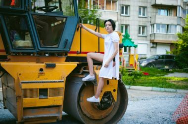 çıplak ayak beyaz spor ayakkabı, yol makine, asfalt finişer ve kentsel manzara fonunda uzun bir forma giymiş olan kadın. Sokak moda.