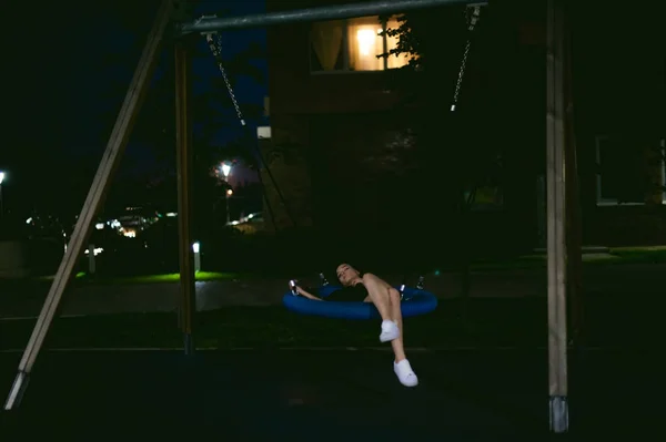 Young woman with beautiful feet dressed in white sneakers and black dress lies in a swing in the yard, against a backdrop of urban landscapes. Street fashion. night dark style — Stock Photo, Image