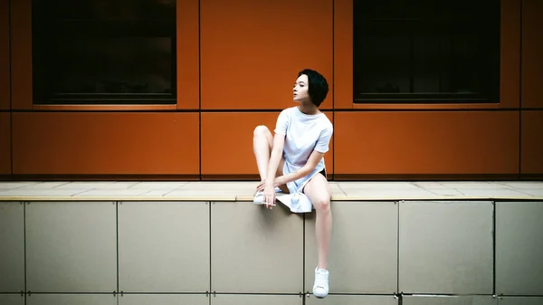 Jeune femme aux pieds nus vêtue de baskets blanches et d'un long T-shirt, fume une cigarette sur fond de paysages urbains. Mode de rue — Photo